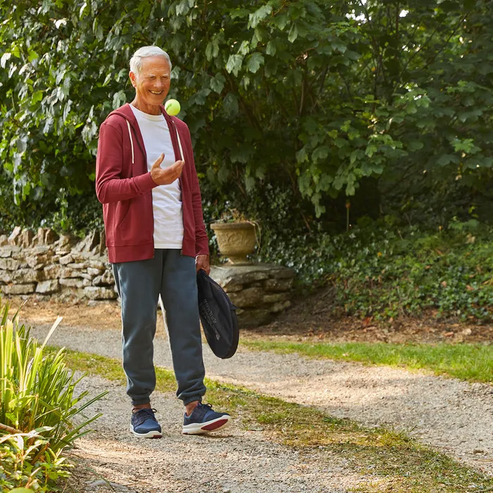Elderly man in a red hoodie and Cosyfeet Vasco Navy, stands on a garden path, smiling while tossing a tennis ball. He holds a hat, with lush greenery and a stone wall behind.