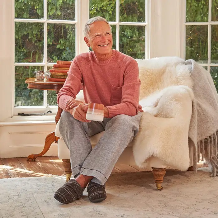 Elderly man in pink sweater and gray pants sits smiling in a Cosyfeet Rudolph Brown Glencoe Slippers sitting in a chair with a glass, by large windows and greenery. Warm and relaxed atmosphere.