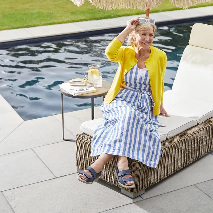 Elderly woman in a yellow cardigan, striped dress and Cosyfeet Sunny in Sapphire Blue relaxes on a wicker lounge chair by a pool, holding sunglasses. A table with lemonade is nearby.