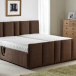A modern bedroom features a large brown upholstered bed with a white mattress, a wooden nightstand with a lamp, a chest of drawers, and a framed picture on the wall.