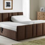 A modern bedroom features a large brown upholstered bed with a white mattress, a wooden nightstand with a lamp, a chest of drawers, and a framed picture on the wall.