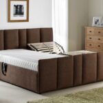 A modern bedroom features a large brown upholstered bed with a white mattress, a wooden nightstand with a lamp, a chest of drawers, and a framed picture on the wall.