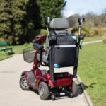 Splash Crutch/Walking Stick Bags Black on a mobility scooter, sitting on a path between two patches of grass in a park environment,