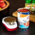 Drive Automatic Can Opener sitting on a kitchen side with part of a can lying on it, open next to the can opener is a open tin of tomatoes, behind a chopping board with herbs and species and a bowl of tomatoes.