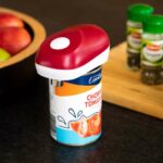 Drive Automatic Can Opener sitting on a tin of tomatoes, behind a bowl of tomatoes and a chopping board holding various herbs and spices.
