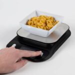 A hand pressing a button the the Drive Digital Talking Scales while a small white bowl sits on top with dry pasta within.