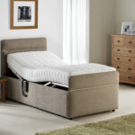 A beige upholstered adjustable bed in a bright room with a raised headrest. Nearby are a wooden nightstand with a lamp, a dresser, and a green rug, creating a cozy, serene atmosphere.