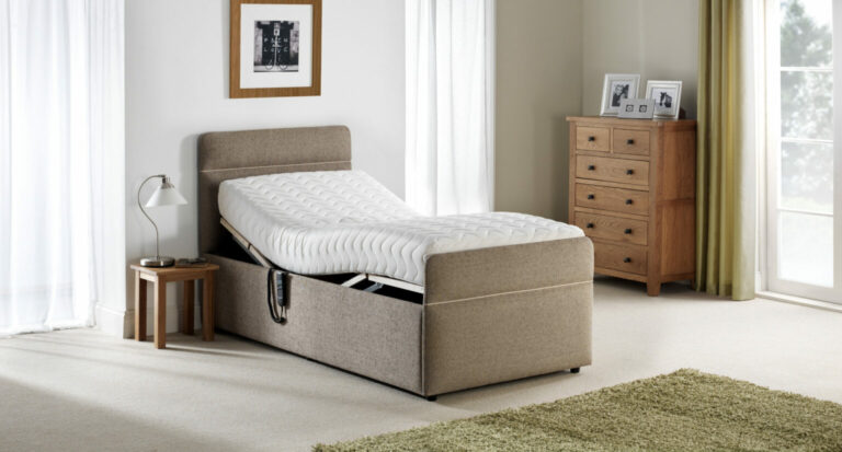 A beige upholstered adjustable bed in a bright room with a raised headrest. Nearby are a wooden nightstand with a lamp, a dresser, and a green rug, creating a cozy, serene atmosphere.