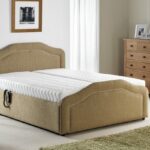 A cosy bedroom with a beige upholstered bed, white mattress, and a side table with a lamp. There's a wooden dresser with photos and a large window with curtains, creating a serene and tidy atmosphere.