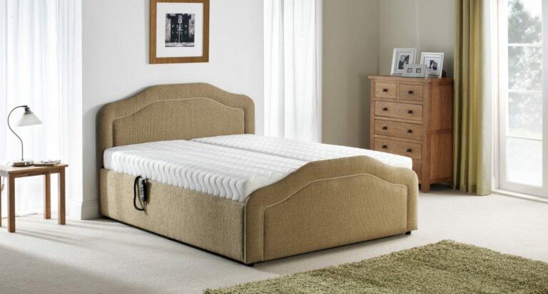 A cosy bedroom with a beige upholstered bed, white mattress, and a side table with a lamp. There's a wooden dresser with photos and a large window with curtains, creating a serene and tidy atmosphere.