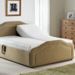 Custom Ollerton Double Bed - A beige adjustable bed with one elevated section is in a bright bedroom. Nearby are a small table with a lamp, and a wooden dresser with pictures.