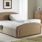 A Custom Selston Single Bed beige adjustable bed with one side elevated in a bright bedroom. Nearby are a wooden table with a lamp and a chest of drawers. The room feels cozy and serene.