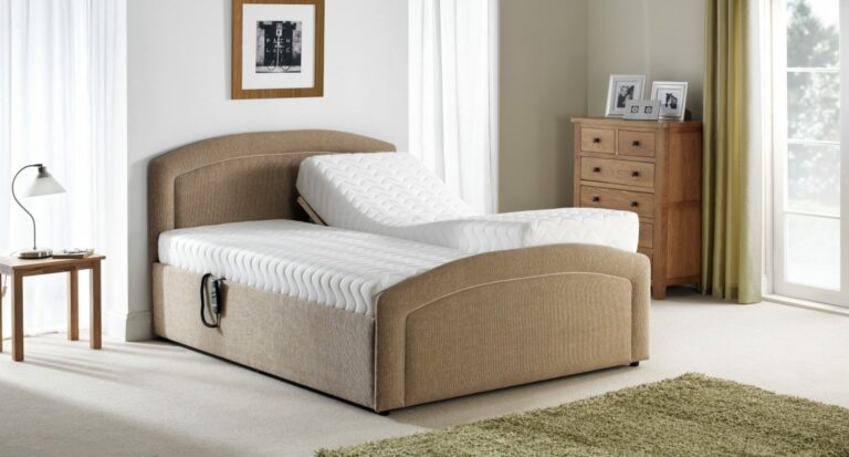 A Custom Selston Single Bed beige adjustable bed with one side elevated in a bright bedroom. Nearby are a wooden table with a lamp and a chest of drawers. The room feels cozy and serene.