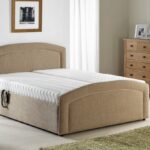 A cosy bedroom with a Custom Selston Double Bed beige upholstered bed and white mattress, a wooden nightstand with a lamp, a dresser with photos, a green rug, and soft natural light.