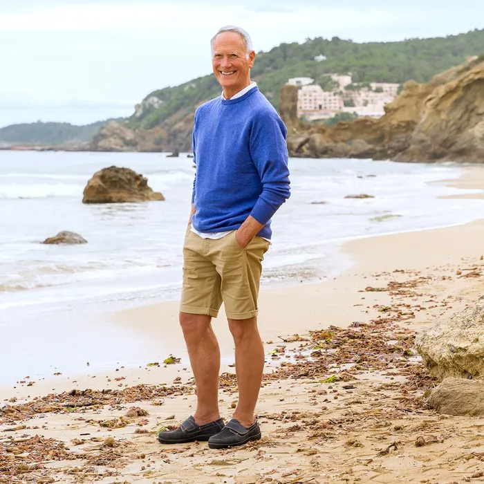 A smiling man in a blue sweater. beige shorts and Cosyfeet Woody Navy boat shoes stands on a sandy beach with rocky cliffs in the background, exuding a relaxed, cheerful vibe.