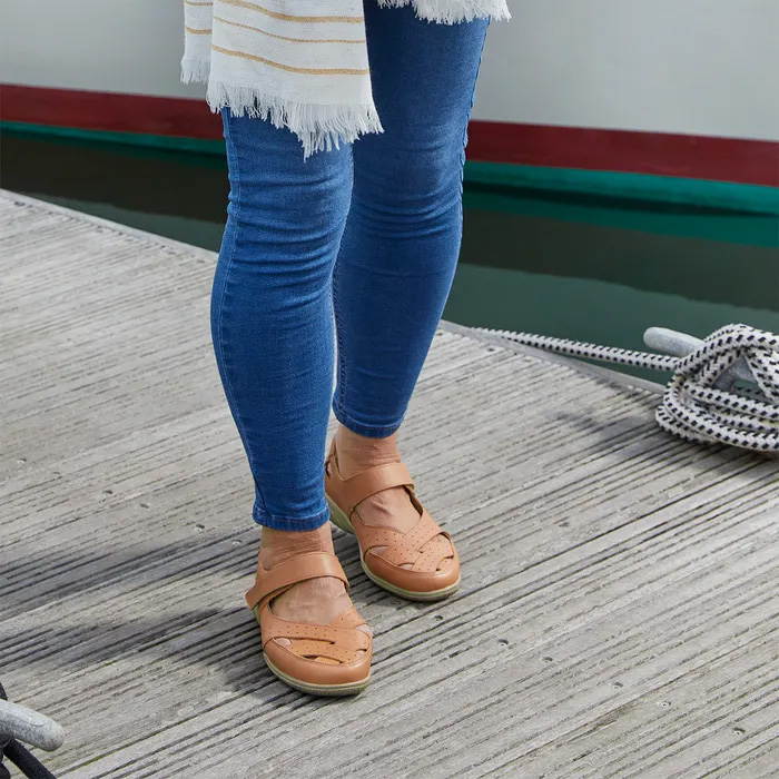 Woman in blue jeans and Cosyfeet Shelley Tan Sandals stands on a wooden dock. A striped shawl drapes over her shoulder, with calm water beside her. Casual, relaxed vibe.