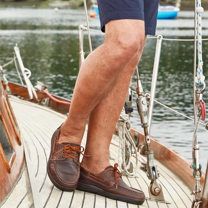 Man Cosyfeets Will Chustnut Brown boat shoes and navy shorts stands on a sailboat deck, with water and blurred shoreline in the background, conveying leisure.