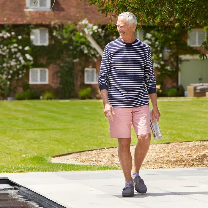 A smiling man with gray hair walks on a patio, wearing a striped shirt, pink shorts and Cosyfeet Woody in Navy, holding a newspaper. Green lawn and a vine-covered house in the background.