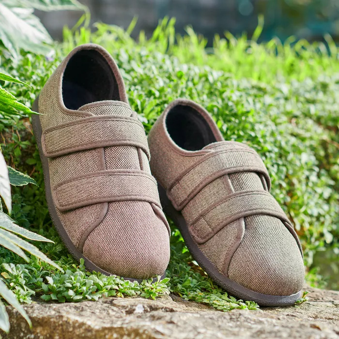 Cosyfeet Steven in Bark with Velcro straps are placed on vibrant green foliage, conveying a natural, rustic feel in an outdoor setting.