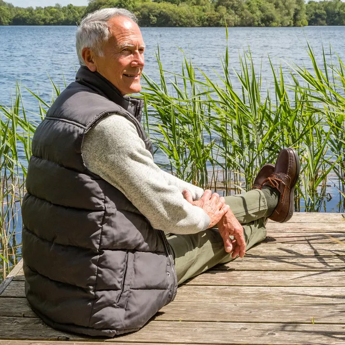 An older man sits on a wooden dock by a lake, wearing a gray puffer vest. green pants and Cosyfeet Will in Chestnut brown. He looks relaxed, surrounded by tall grass, with trees in the background.