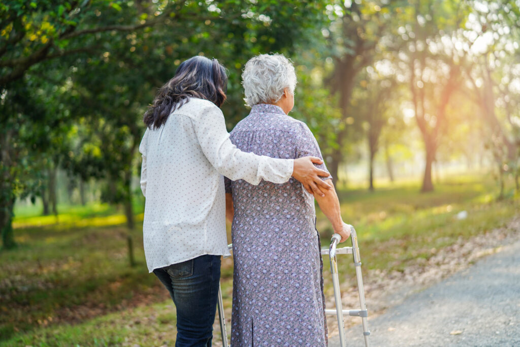 A woman supporting an elderly woman as she walks through a park with her walking frame.