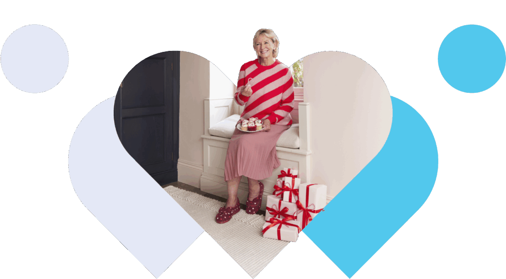 A woman in a red and white striped sweater and pink skirt, wearing Cosyfeet slippers on, sits on a white bench, smiling. She holds a mug beside wrapped gifts. Cosy and festive ambiance.