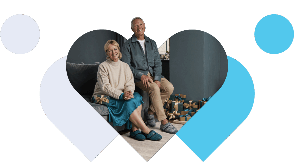 An elderly couple sits smiling on a sofa, wearing Cosyfeet Slippers surrounded by wrapped gifts. They're framed by a heart shape, creating a warm, joyful atmosphere.