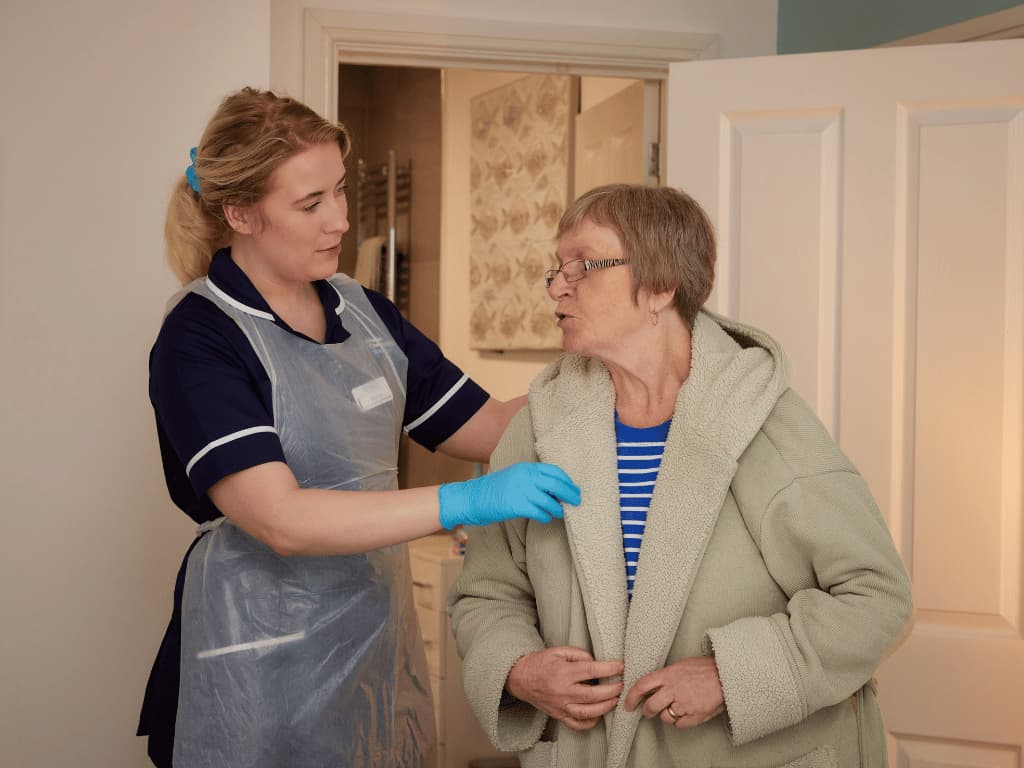 A carer wearing a dark blue uniform is helping a lady put on a dressing gown to represent Live-In Respite Care at Premier Community.