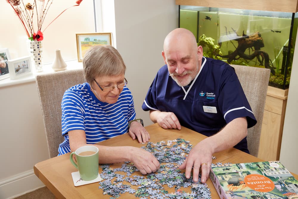 A male carer sits with a female service user and is helping to complete a puzzle to reflect person-centred care for those with dementia
