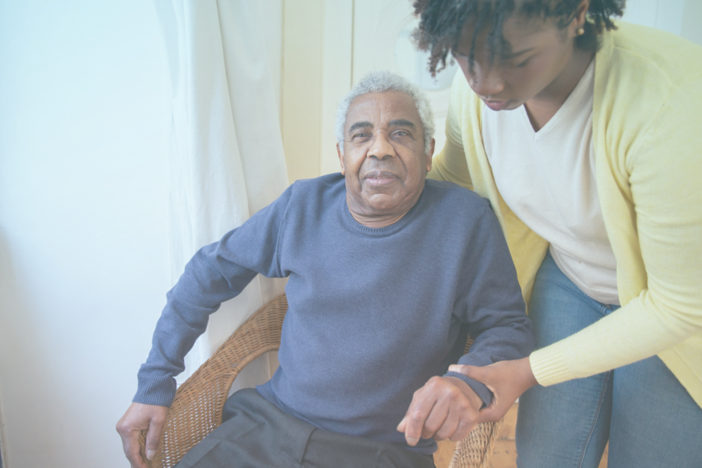 An elderly man with a blue jumper sitting in a chair while a lady in a white top and yellow cardigan hold his arm, bending over to assist.