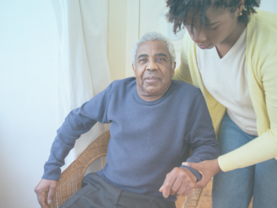 An elderly man with a blue jumper sitting in a chair while a lady in a white top and yellow cardigan hold his arm, bending over to assist.