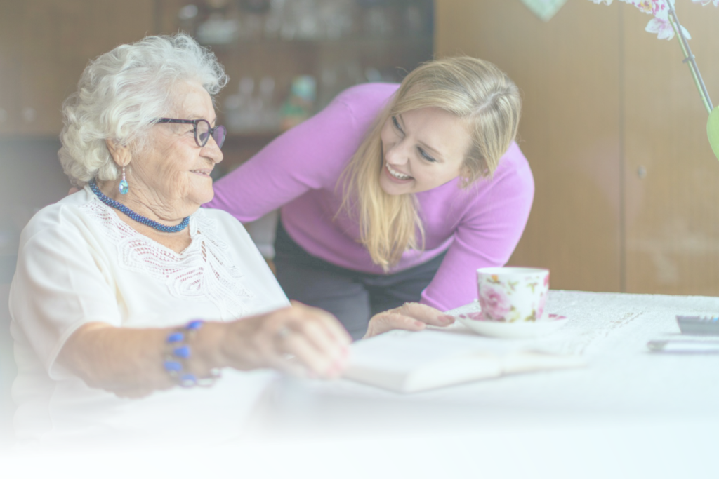 An elderly lady in a white top holding a book, while a younger blonde lady in a purple top bends down to talk to her.