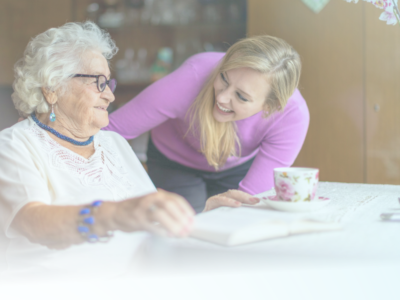 An elderly lady in a white top holding a book, while a younger blonde lady in a purple top bends down to talk to her.