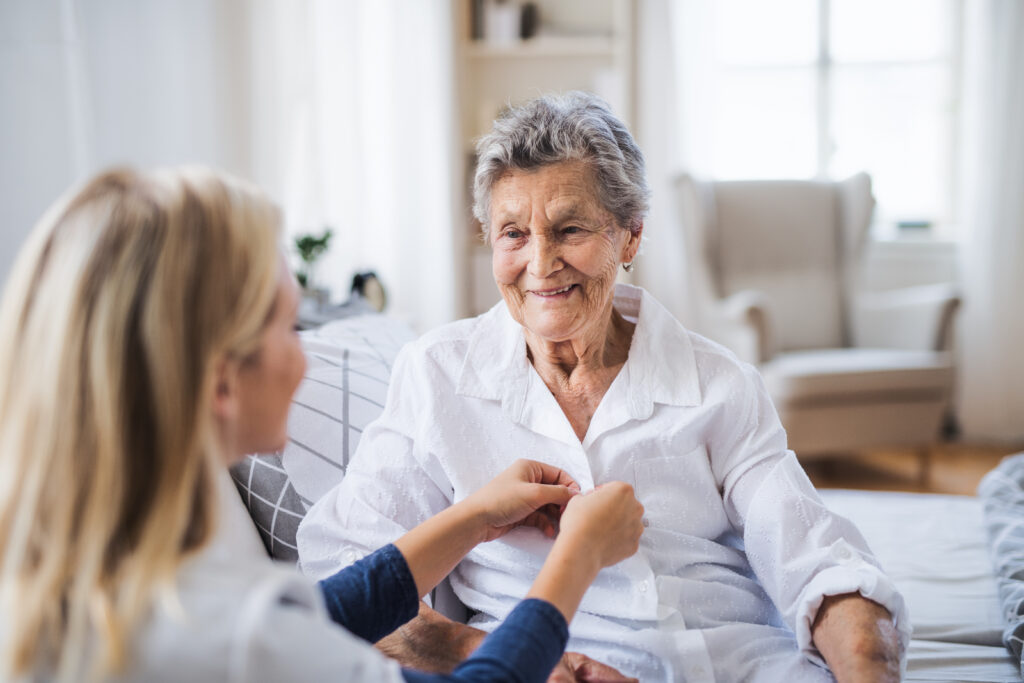 A young visitor helping a happy sick senior woman sitting on bed at home.
