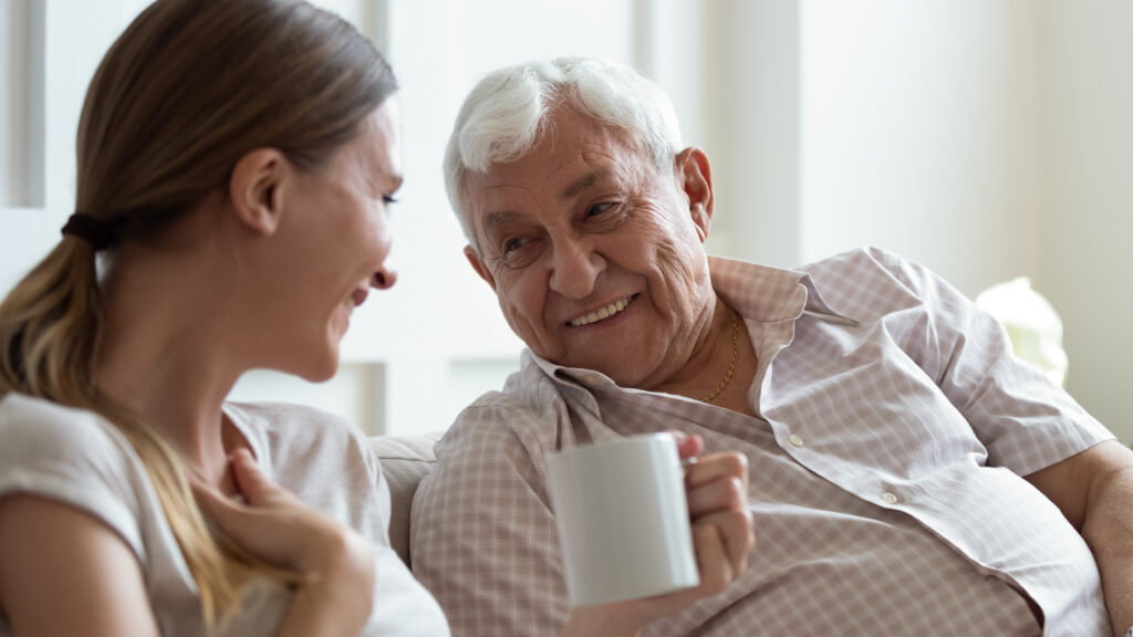 Close up smiling older father and adult daughter chatting, drinking tea or coffee, sitting on cozy couch at home, positive mature grandfather and young woman having fun, talking, sharing news