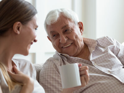Close up smiling older father and adult daughter chatting, drinking tea or coffee, sitting on cozy couch at home, positive mature grandfather and young woman having fun, talking, sharing news